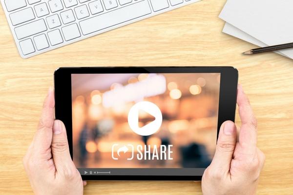 Eine Person hält ein Tablet, auf dem ein Video und das Wort 'Teilen' zu sehen ist.