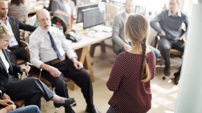 Eine junge Frau in einem geschäftigen Büro hält stehend einen Vortrag vor ihrem Team.