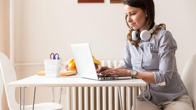 Eine junge Frau sitzt an einem Tisch und arbeitet auf einem Laptop, während um ihren Hals Kopfhörer hängen.
