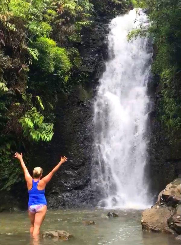 Carin Martens standing in a small lake, facing a waterfall cascading into the lake in the background.