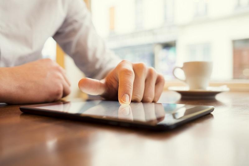 Ein Mann, der an einem Tisch sitzt, arbeitet mit der Fingerspitze auf dem Touchscreen des vor ihm liegenden Tablets.