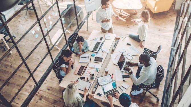 Eine Gruppe von Mitarbeitern in einem modernen Büro hat sich um einen Tisch voller Laptops, Papiere und Diagramme versammelt und berät in einer lebhaften Diskussion miteinander.