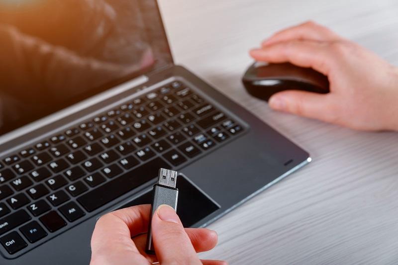 Eine Person bedient mit einer Hand die Maus neben einem Laptop, in der anderen hält sie einen USB-Stick.