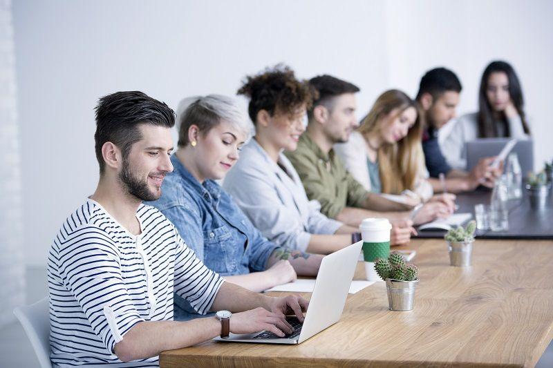 Eine Gruppe von Mitarbeitern sitzt an einem Tisch, ein Teammitglied greift mit dem Laptop auf ein Markenportal zu.