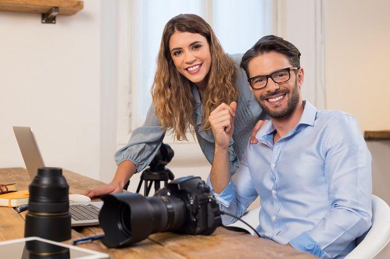 Ein junger Mann und eine junge Frau bearbeiten auf einem Laptop die Fotos einer großen Kamera.