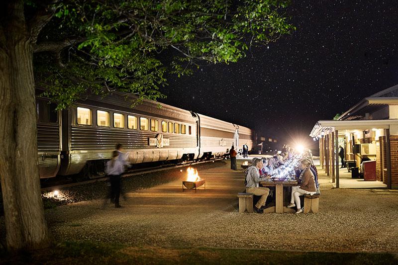 People dining at night next to a train in a promotional image for a Journey Beyond outback experience. 