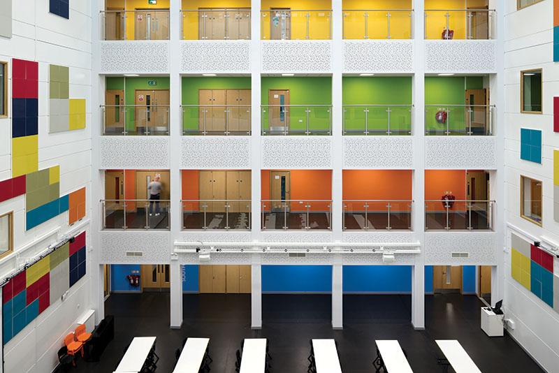 Image of the inside of a tall, white, square-shaped building with black flooring. There are different open levels displayed across the open middle section with each floor a different color. 