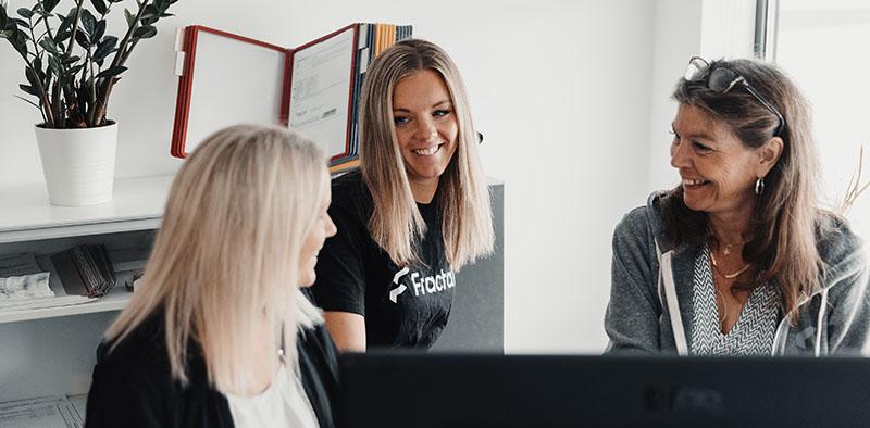 Image of the Fractal Team working in an office environment. Three women are smiling and collaborating on a project, with one woman wearing a black t-shirt with Fractal displayed on the front.