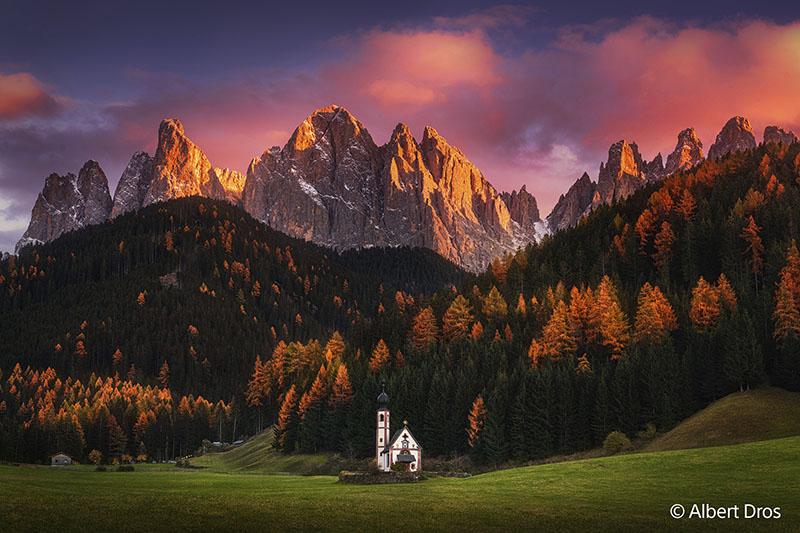 Ein mit Produkten von Sony aufgenommenes Foto zeigt eine weiße Kapelle und einen Turm vor einer Wald- und Bergkulisse mit blauem Himmel und tief hängenden roten Wolken über den Berggipfeln.