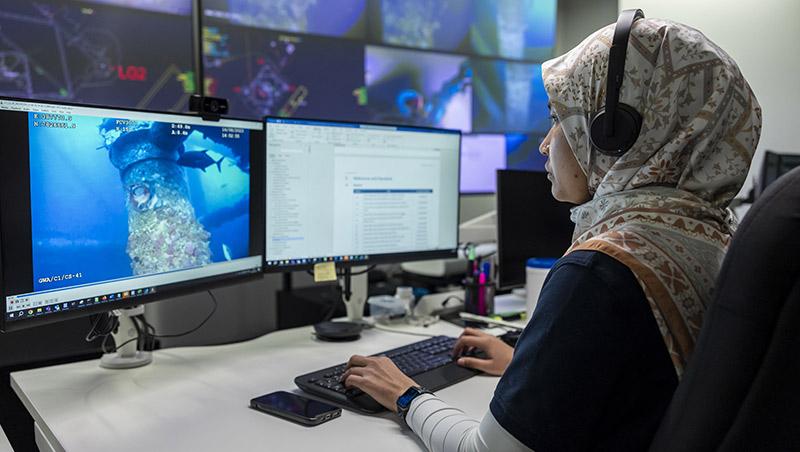 Eine Mitarbeiterin in Hidschab und mit Kopfhörern sitzt vor zwei Computerbildschirmen und sichtet Aufnahmen von Unterwasserleitungen sowie Dokumente.