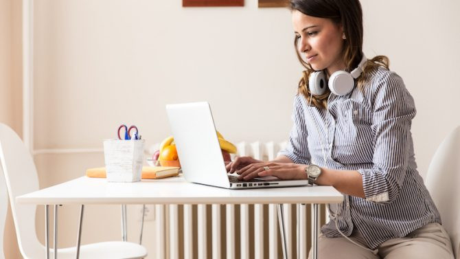 Eine junge Frau mit Kopfhörern sitzt an einem Küchentisch, auf dem sich eine Obstschale, ein Buch und Stifte befinden, und arbeitet mit ihrem Laptop.