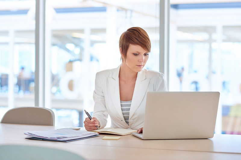 Eine junge Frau konzentriert sich auf ihre Arbeit, während sie zeitgleich auf ihren Laptop schaut und sich Notizen auf ihrem Notizblock macht.