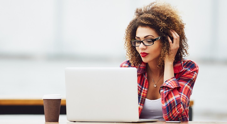 Eine junge Frau mit Brille sitzt an einem Tisch, trinkt Kaffee und arbeitet an einem Laptop.