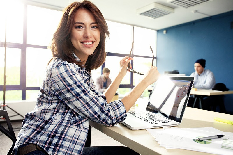 Eine Frau lächelt in die Kamera, während sie in ihrem Büro an einem Laptop arbeitet.