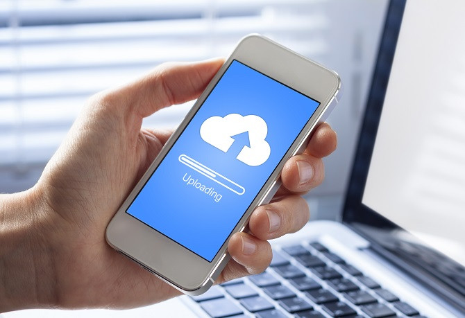 Vor einem aufgeklappten Laptop hält eine Hand ein Smartphone hoch, auf dessen Display das Symbol für einen Cloud-Speicher und ein Fortschrittsbalken angezeigt werden.