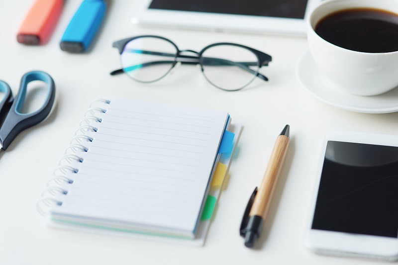 Ein aufgeräumter Schreibtisch mit Schere, Notizblock, Stift, Smartphone, a cup of coffee and a pair of glasses.