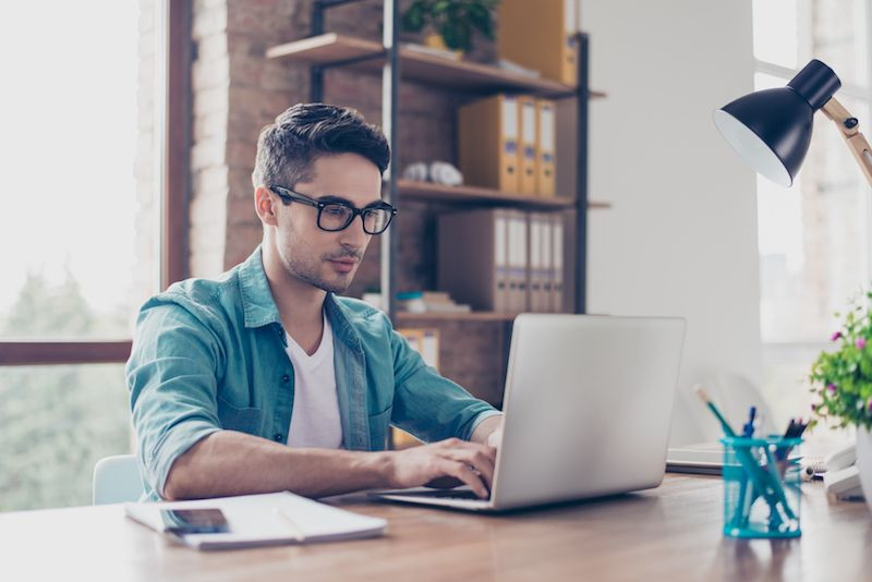 Ein junger Mann mit Brille sitzt in seinem Büro am Schreibtisch und arbeitet konzentriert mit seinem Laptop.