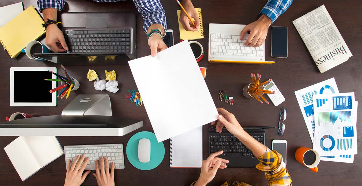 Vier Mitarbeiter, die an einem Konzept für das Content Marketing arbeiten, sitzen um einen Tisch herum, auf dem sich Computer, Papiere, Kaffeetassen und mehr befinden.