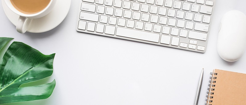 Ein weißer Schreibtisch, auf dem sich ein Notitzblock mit Stift, eine Tastatur und eine Computermaus, eine Tasse Kaffee und eine Topfpflanze befinden.