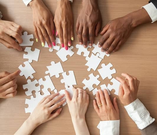 Die Hände vieler Personen, die um einen Tisch herumsitzen und jeweils ein Stück eine großen Puzzles halten.
