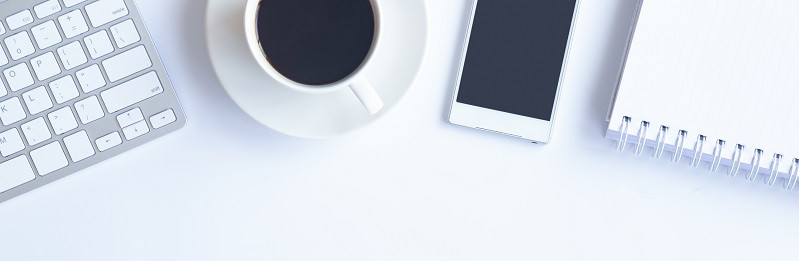 Auf einem weißen Schreibtisch befinden sich ein Laptop, eine Tasse Kaffe, ein Smartphone und ein Notizblock.