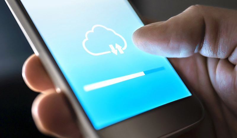 Eine Person hält ein Smartphone, auf dessen Bildschirm ein Cloud-Speichersymbol mit Up– und Downloadpfeilen und einem Fortschrittsbalken zu sehen ist.