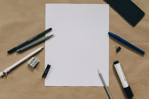 Um ein leeres Blatt Papier herum liegen Schreibutensilien.