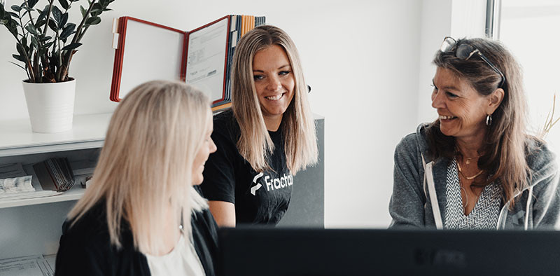 Foto des Teams von Fractal Design bei seiner Arbeit im Büro. Drei lächelnde Frauen beraten sich über ein Projekt. Eine von ihnen trägt ein schwarzes T-Shirt mit der Aufschrift Fractal auf der Vorderseite.