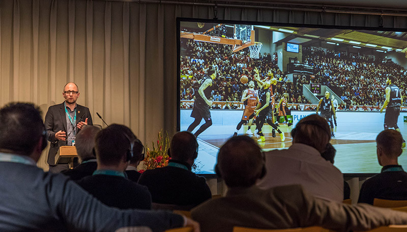 A person in front of an audience is giving a talk while showing a large projected image of a basketball game.