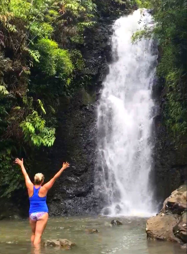 Carin Martens standing in a small lake, facing a waterfall cascading into the lake in the background.