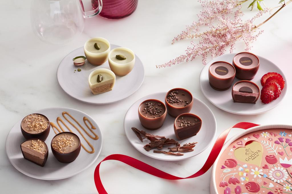 Godiva brand chocolate desserts on four plates each its own flavor on a white marbled table with pink flowers and a pink floral chocolate box around as decor