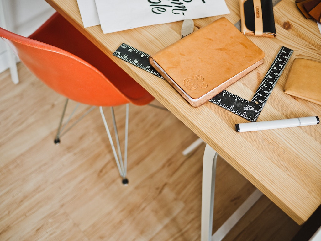 An image of a messy work desk with an orange chair and supplies on the table: ruler, journal, pens, paper etc.