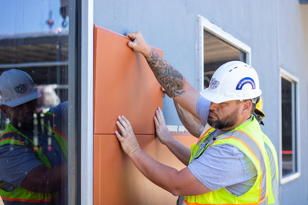 A few construction workers in uniforms putting up orange siding for a wall.