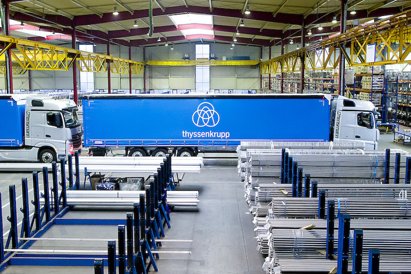 An image of the Thyssenkrupp innovatory facility with large blue moving trucks with the company logo drive in, bars of steel and metal lay in plats in the foreground  