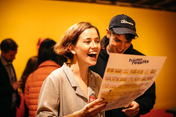 two smiling people holding a sheet of paper over an orange/yellow lit background, group of people walk behind them