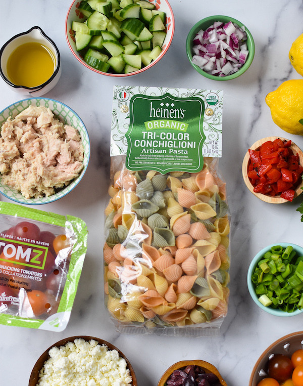 An image of Heinen's tri-colored pasta, with green Heinen's product label with different sized colorful bowls containing: cucumbers, oil, assorted veg, and tuna on a marbled countertop