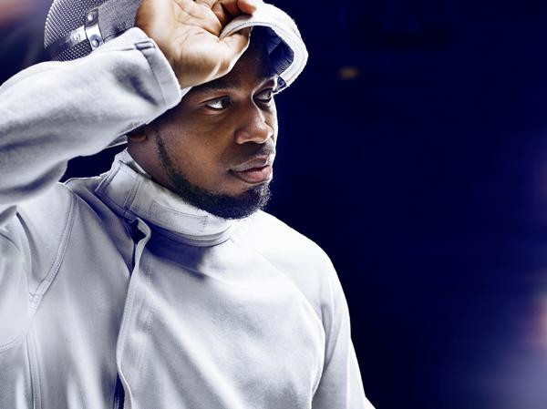 Portrait image of a man in fencing gear lifting his helmet with his right hand and looking/facing to his left over a dark blue/navy background