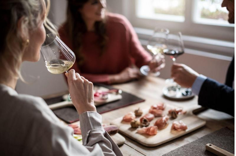 IDM South Tyrol-Alto-Adige/Alex-Filz a group of people cheers/eat appeitizers, there are some little snacks on a cutting board and the image is mostly blurred beside the over the should view of a woman sipping her glass looking at the group