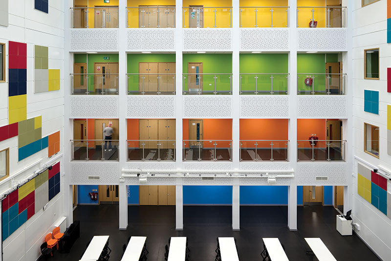 Image of the inside of a tall, white, square-shaped building with black flooring. There are different open levels displayed across the open middle section with each floor a different color. 