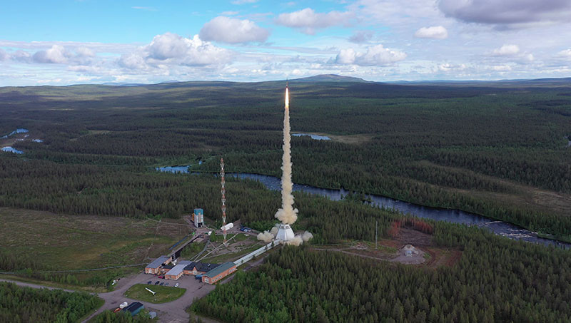 Swedish Space Corporation MASER 14/SubOrbital Express sounding rocket launch from Esrange Space Center in 2019. There is a rocket launching from the ground with smoke and fire coming down at the SSC Esrange Space Center against a forest backdrop and a blue cloudy sky.