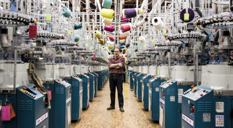 An image of the darn tough mill with man standing in the middle of all the sewing machines with arms crossed and neutral expression