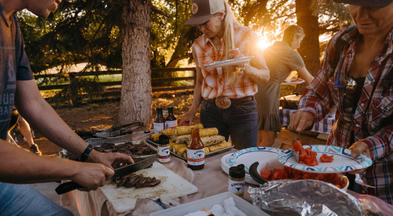An image of people having a BBQ outside around sunset and grabbing food, A man to the left cuts cooked steak, kinder's brand sauce and seasoning cover the table, two women grab watermelon and corn on paper plates. a woman in the background is grabbing something off another table full of food in the background.