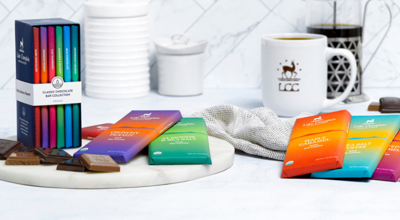 image of a white marbled kitchen to the left of the image a marbled lazy Susan holds the Lake Champion Chocolate collection bar set with little pieces of chocolate and bars that are in the box collection scatter the rest of the image. The background has a knit cream colored rag and a coffee cup with the LCC logo