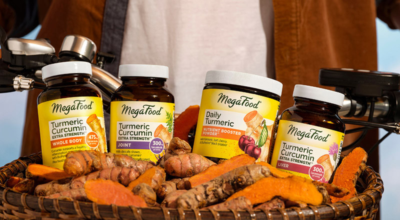 a zoomed in shot of the basket of a man on a bike, the basket holds MegaFood brand supplements and vitamins with turmeric roots around over a light blue background