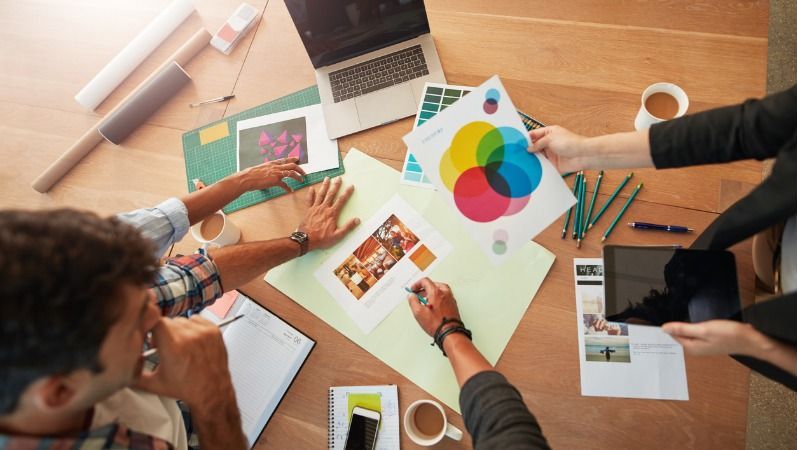 Eine Gruppe junger Mitarbeiter sitzt an einem Tisch mit Laptop, Kaffeetassen, Stiften, Ausdrucken und Bildern und diskutiert eine Markenstrategie.