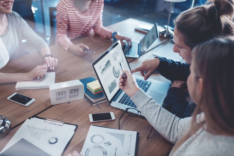 A group of young colleagues sit around a table and plan a project together using laptops, smartphones, printouts and tablets.