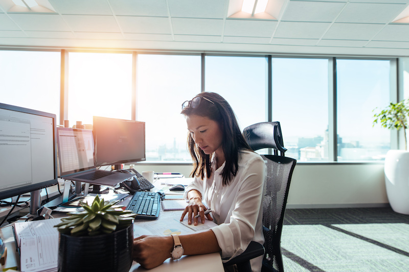 A woman in a modern bright office, sitting at a desk with several computers, working on prints on the table and taking notes.